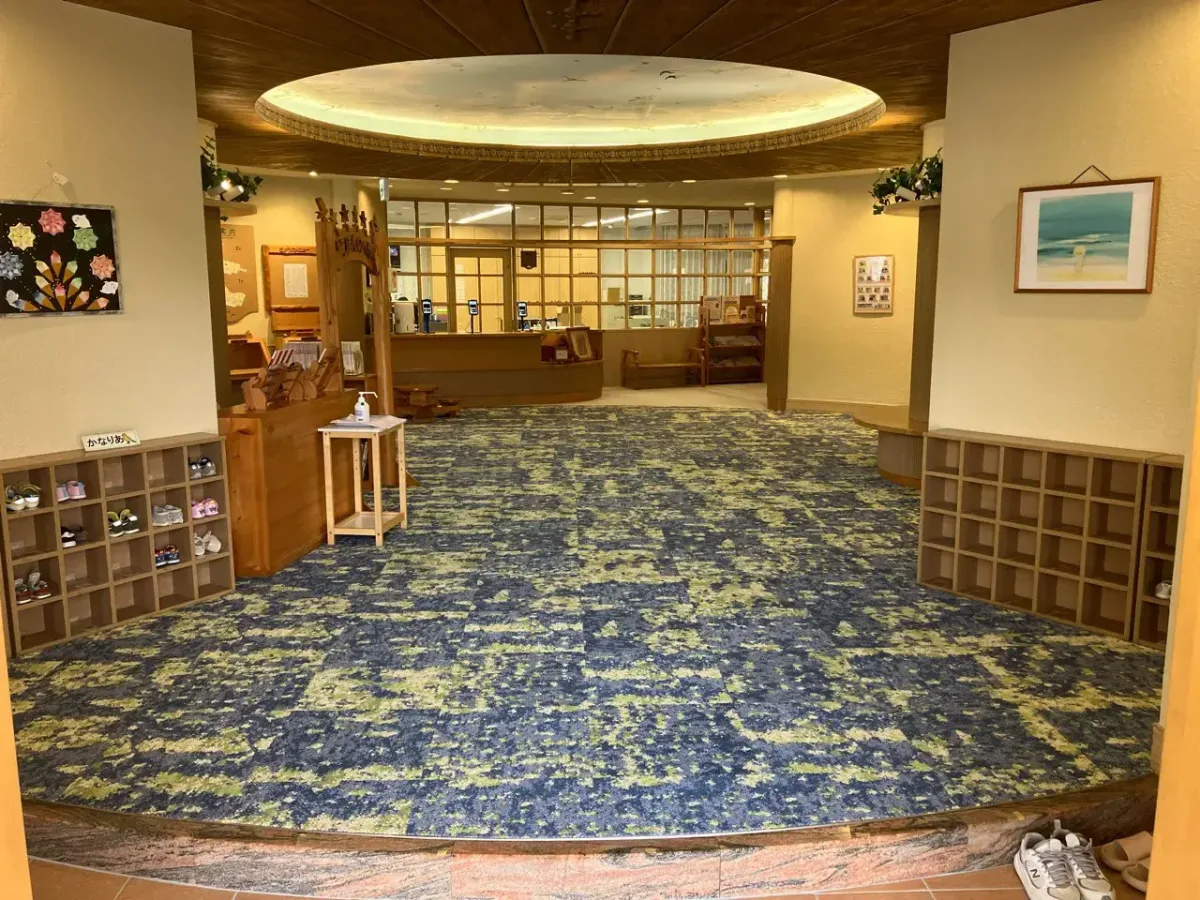 モネの「睡蓮の池」をイメージしたという施設のエントランスホール。床には青と緑が混ざり合った印象派風のカーペットが敷かれ、天井には円形の大きな照明があります。左右には木製の下駄箱があり、奥には受付カウンターが見える広々とした空間です。