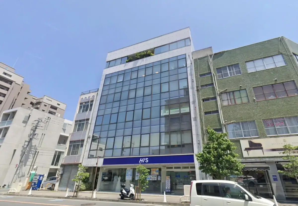 一栋玻璃幕墙中层大楼的外观。一楼设有HIS门店，前方道路上停着一辆白色轿车和一辆摩托车。这张街景照片拍摄于晴朗的蓝天之下，两旁排列着设计各异的大楼。